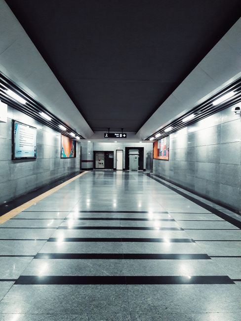 View of an underground railway station platform seen through an oval-shaped window, showing clean wooden wall panels, a tiled floor, and a glass door with a metallic frame. The station features modern